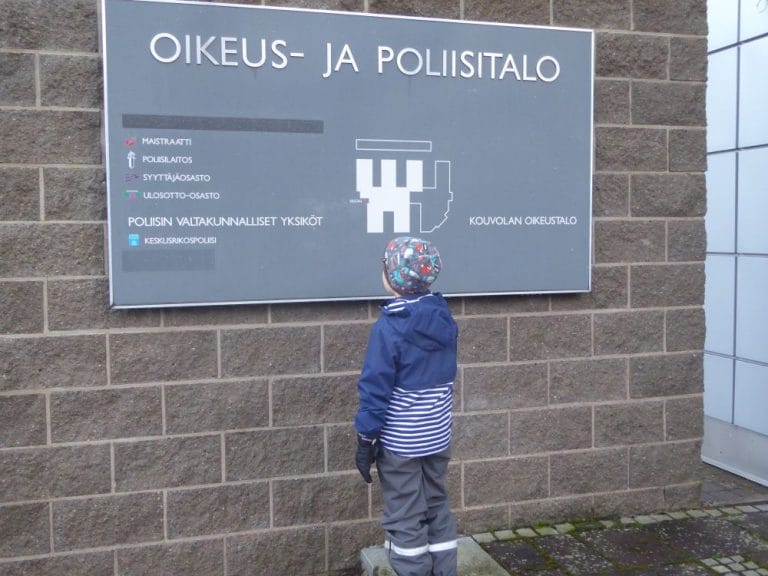 Lapsi seisoo selin kameraan oikeus- ja poliisitalon opastekyltin edessä.