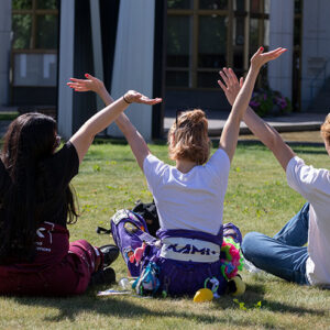 Kolme opiskelijaa istuu selin nurmikolla kädet taivasta kohti.