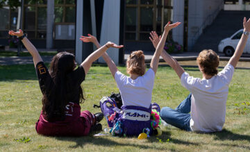 Kolme opiskelijaa istuu selin nurmikolla kädet taivasta kohti.