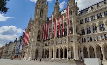 Kuvassa näkyy Wienin kaupungintalo, joka muistuttaa goottilaisia katedraaleja ja rakennuksen etuyläosaan on kiinnitetty punavalkoisia pitkiä viirejä.